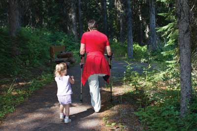 Photo of a little girl and a man, who is using crutches, walking on a trail in a forested area.