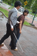 Photo of a man who is blind taking the elbow of a friend to help him navigate a curb and walkway.