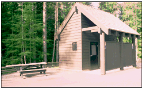 A photograph of a public toilet building in a wooded park. An accessible entrance is provided to the toilet building. An accessible picnic table is shown to the left of the building.