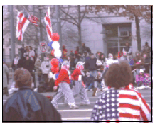 A photograph of costumed people walking down a parade route, with spectators on either side of the street.