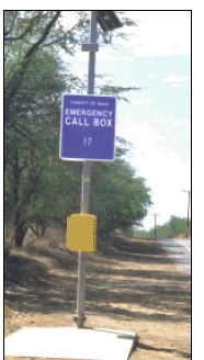 A photograph of a roadside emergency call station along a two-lane rural highway.