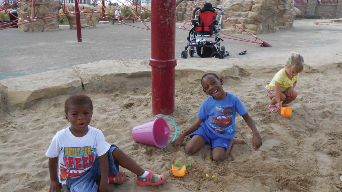 Two pre-school age brothers play in the sand with a little girl.  A wheelchair is situated behind them.