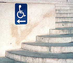 A photograph of stairs with a blue and white directional sign with the international symbol of accessibility pointing toward the left in the direction of the accessible entrance.
