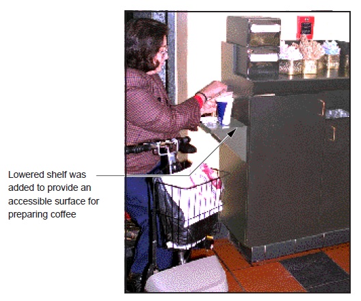 photo - person using a scooter pulled alongside a shelf that has been installed to provide accessible condiments. note: Lowered shelf was added to provide an accessible surface for preparing coffee.