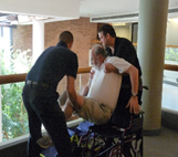 Two men transfering another man from his wheelchair into and "emergency stair desecent device."