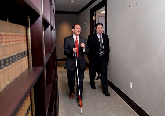 Two workers, one of whom is visually impaired and uses a cane, walking in an office.